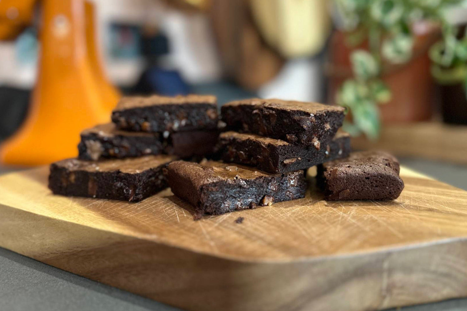 A stack of rich, fudgy chocolate brownies with a glossy top and chunks of chocolate, displayed on a wooden board in a home kitchen setting